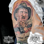 Home new Highly detailed color realism horror half-sleeve tattoo on a man's upper arm and shoulder by Mad Time Tattoo, featuring a portrait of Freddy Krueger with his iconic razor-glove, blended with a scene of little girls skipping rope outside the Elm Street house below.