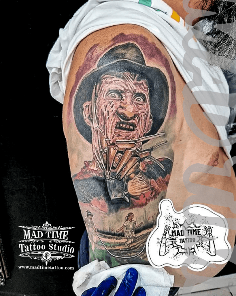 colour-realism-tattoos-liverpool Highly detailed color realism horror half-sleeve tattoo on a man's upper arm and shoulder by Mad Time Tattoo, featuring a portrait of Freddy Krueger with his iconic razor-glove, blended with a scene of little girls skipping rope outside the Elm Street house below.