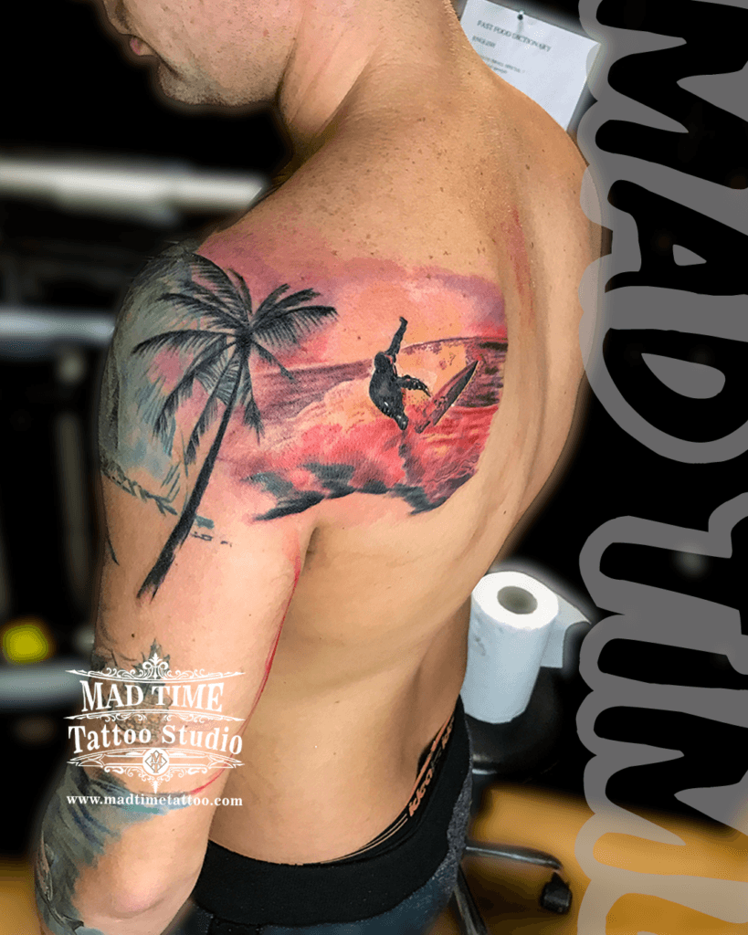 colour-realism-tattoos-liverpool Stunning color realism landscape tattoo spanning the shoulder blade and upper arm, depicting a silhouette surfer riding a big wave under a vibrant pink and orange sunset sky with a detailed palm tree by Mad Time Tattoo.