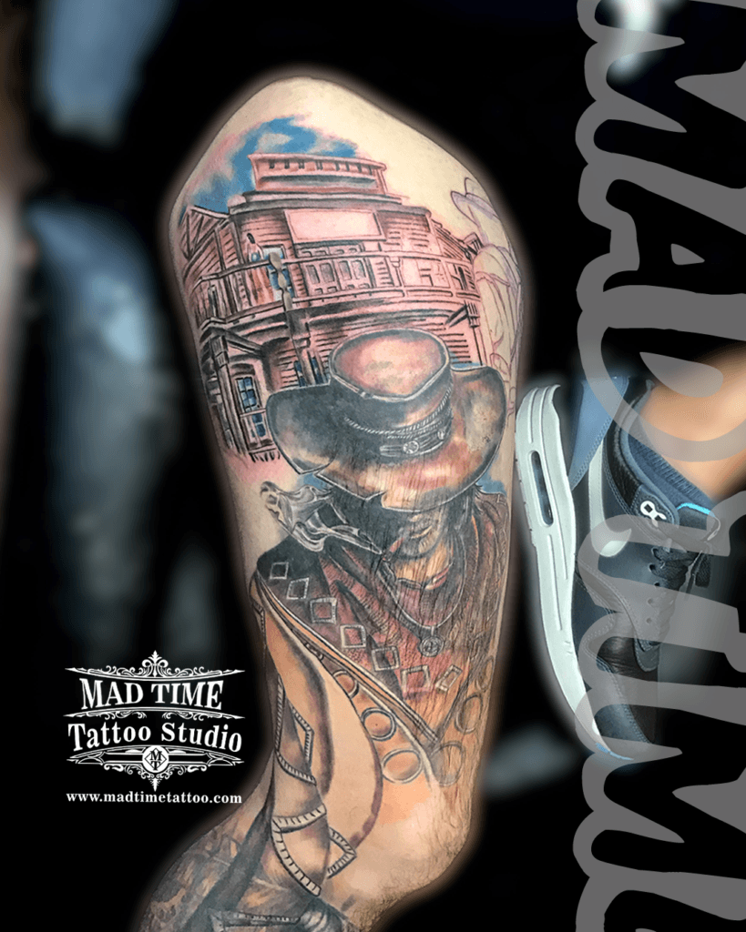 colour-realism-tattoos-liverpool Illustrative black and grey tattoo with color accents on an upper arm, depicting a smoking cowboy wearing a leather hat and poncho in front of a rustic Wild West wooden saloon building by Mad Time Tattoo