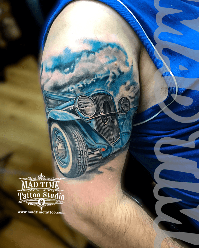 colour-realism-tattoos-liverpool Close-up color realism tattoo of a classic vintage blue car on a man's upper arm by Mad Time Tattoo, showcasing detailed chrome headlights, textured tires, and a scenic cloudy blue sky backdrop.