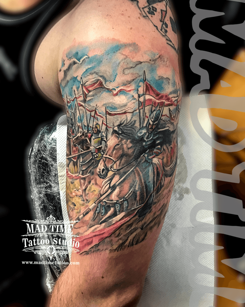 colour-realism-tattoos-liverpool Dynamic color and blackwork realism tattoo of a Polish Hussar cavalry knight charging on a horse with a lance, long spears, and red and white flags under a cloudy sky on a man's upper arm by Mad Time Tattoo.