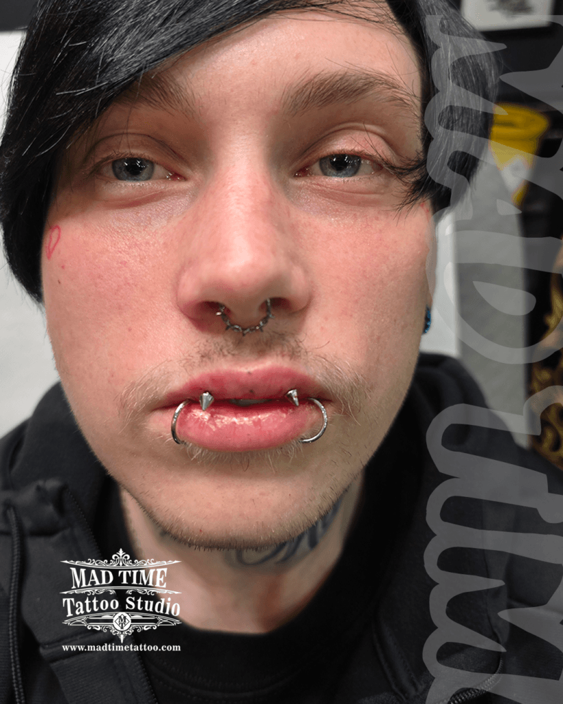 Edgy custom lip piercing setup at Mad Time Tattoo Studio, featuring lower lip snake bites styled with large titanium captive bead rings and sharp upper cone spikes, paired with a textured septum hoop.