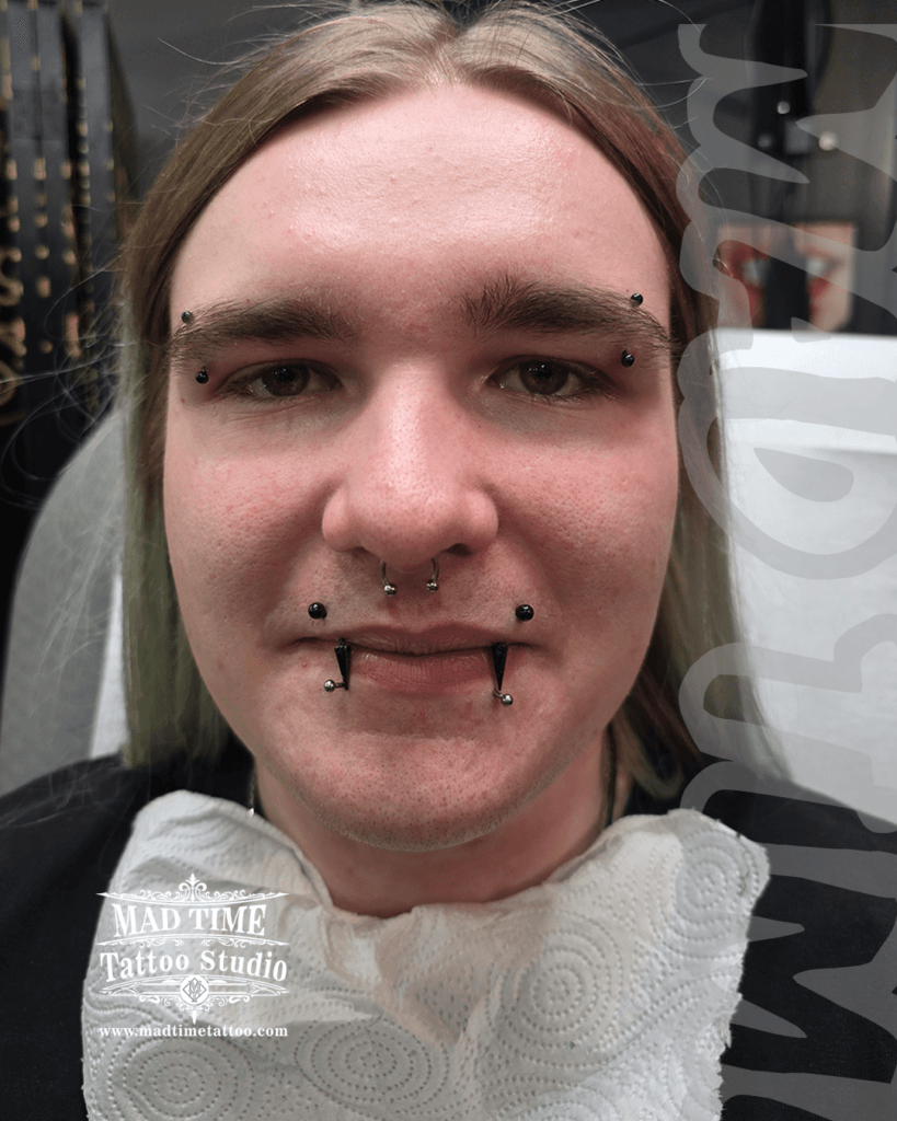 Advanced facial piercing curation by Mad Time Tattoo Studio, showcasing symmetrical snake bites with black spike jewelry, paired with matching double eyebrow piercings, angel bites, and a septum hoop.