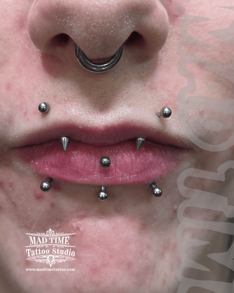 Close-up of an advanced oral piercing curation at Mad Time Tattoo Studio, featuring a stacked combination of snake bites with lower inner spikes, angel bites, a vertical labret, and a septum clicker hoop.