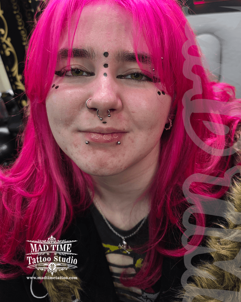 Full face alternative piercing layout showcase by Mad Time Tattoo Studio on a client with vibrant pink hair, highlighting snake bites studs, a medusa stud, nostril hoop, septum ring, and vertical bridge dermal studs.