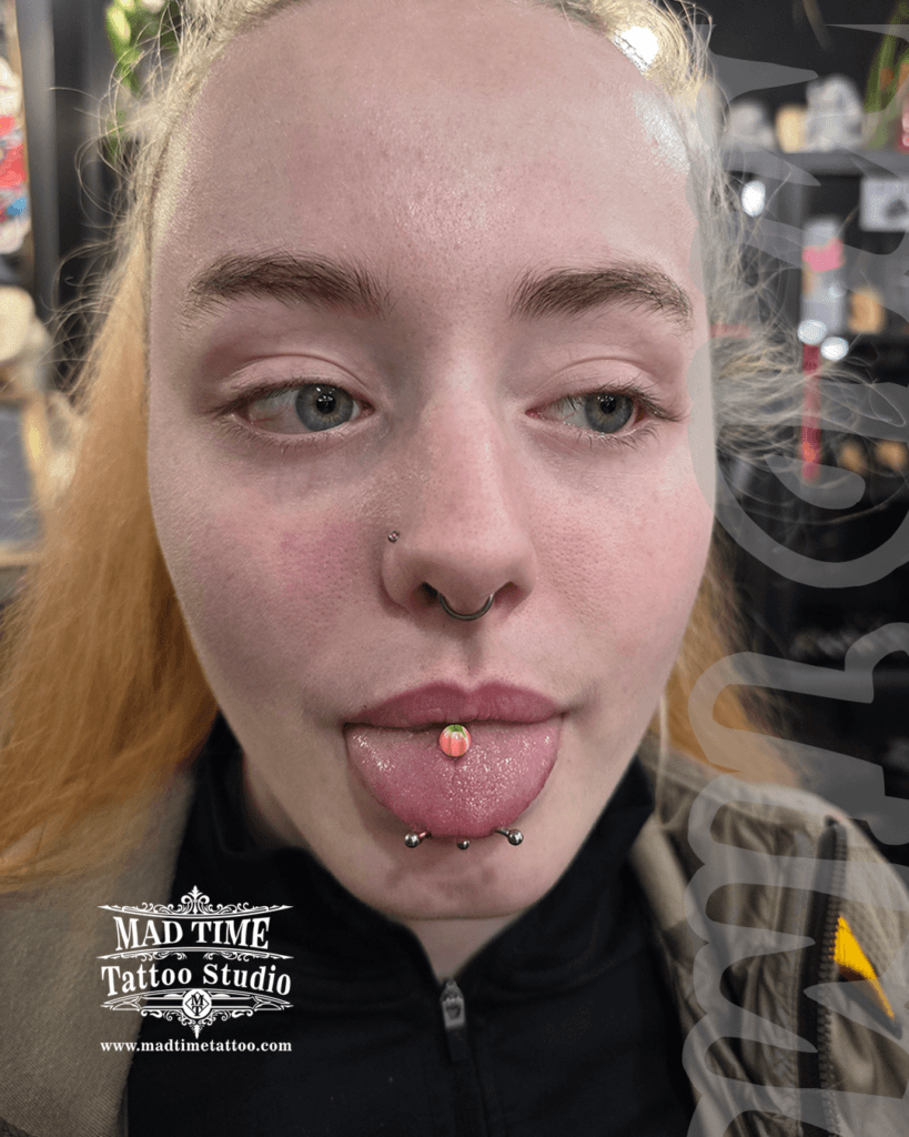 • Full face body piercing curation by Mad Time Tattoo Studio, showcasing a horizontal snake eyes piercing on the tongue tip, stacked behind a central midline tongue bar with a pink opal gem top, a black septum ring, and a nostril stud.