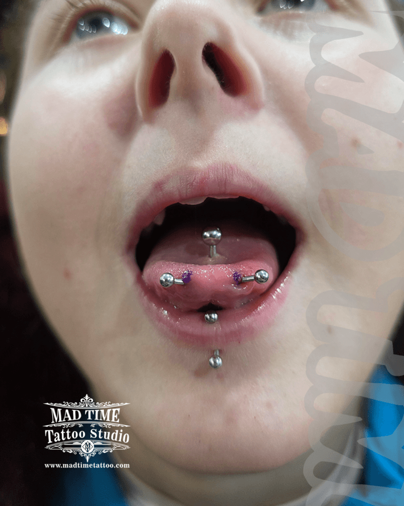 Macro view of an advanced oral modification setup by Mad Time Tattoo Studio, showcasing a horizontal snake eyes tongue piercing with surgical markings, a classic midline tongue barbell, and a vertical labret piercing on the lower lip.