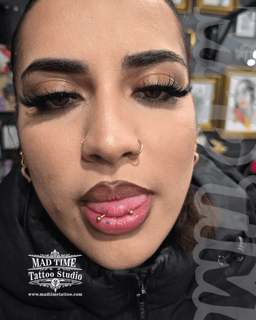 • Professional snake eyes tongue piercing showcase at Mad Time Tattoo Studio, featuring a horizontal tongue tip barbell with premium gold ball configurations, paired with matching gold double nostril hoops.