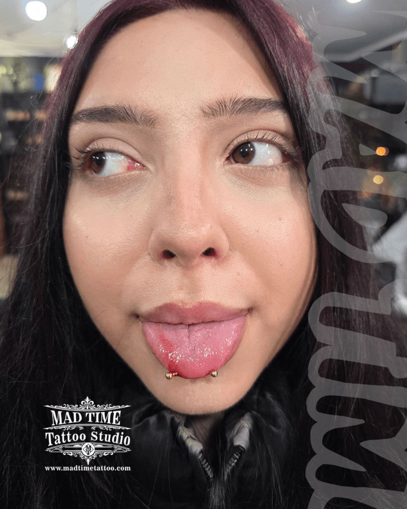 • Close-up of a fresh horizontal snake eyes tongue piercing at Mad Time Tattoo Studio, featuring perfectly balanced gold-anodized titanium beads on a female client.