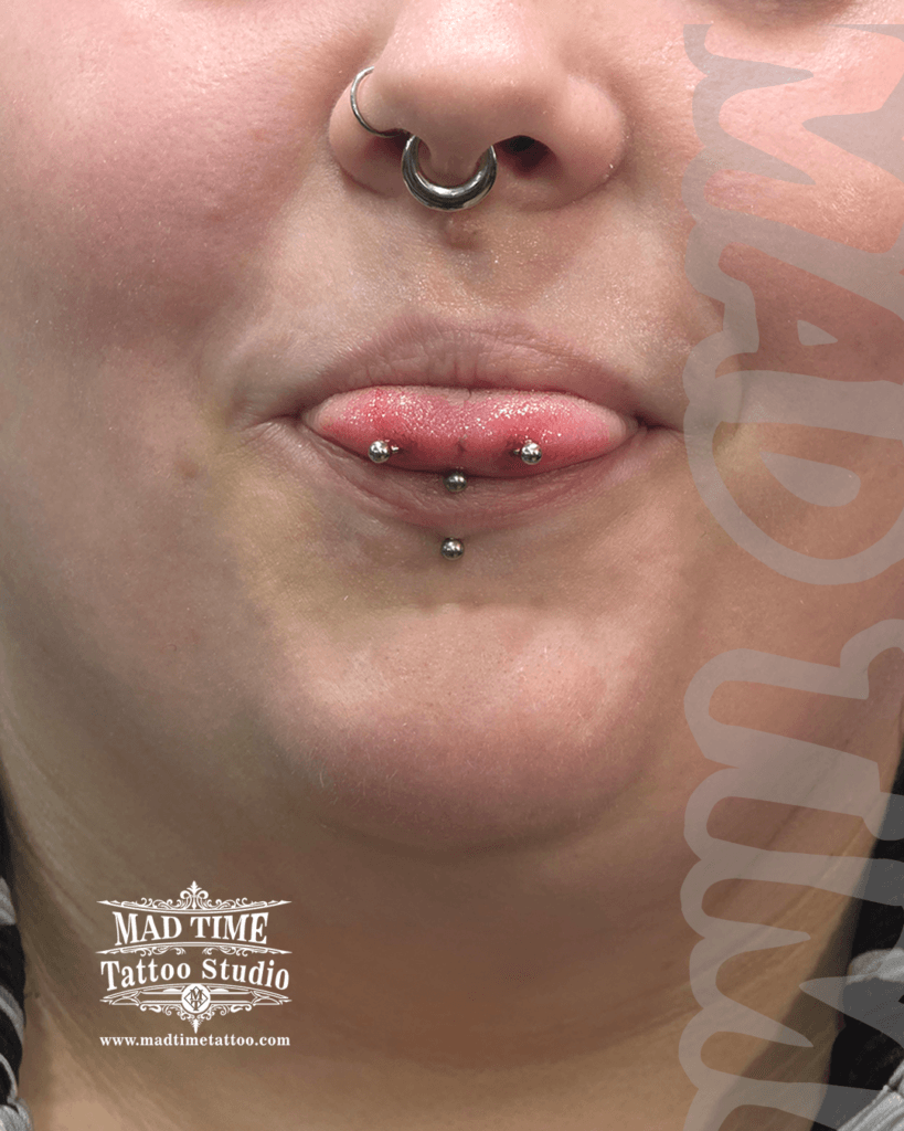 Macro view of an advanced oral modification stack at Mad Time Tattoo Studio, highlighting a horizontal silver snake eyes piercing, traditional midline tongue bar, and lower lip vertical labret.