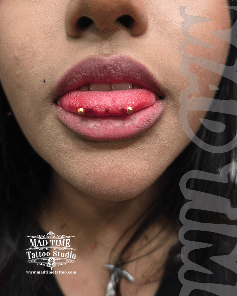 High-precision horizontal snake eyes tongue piercing at Mad Time Tattoo Studio, styled with vibrant gold-anodized titanium curved barbell beads at the tip of the lips.