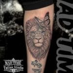 Home new Highly detailed black and grey realism tattoo of a lion's head with bright blue eyes and a fine-line butterfly on its forehead, finished with custom names Ruby and Bella on a lower leg or arm by Mad Time Tattoo.
