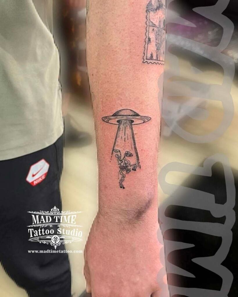 Minimalist blackwork tattoo depicting a small UFO beam abducting something, placed on a forearm.