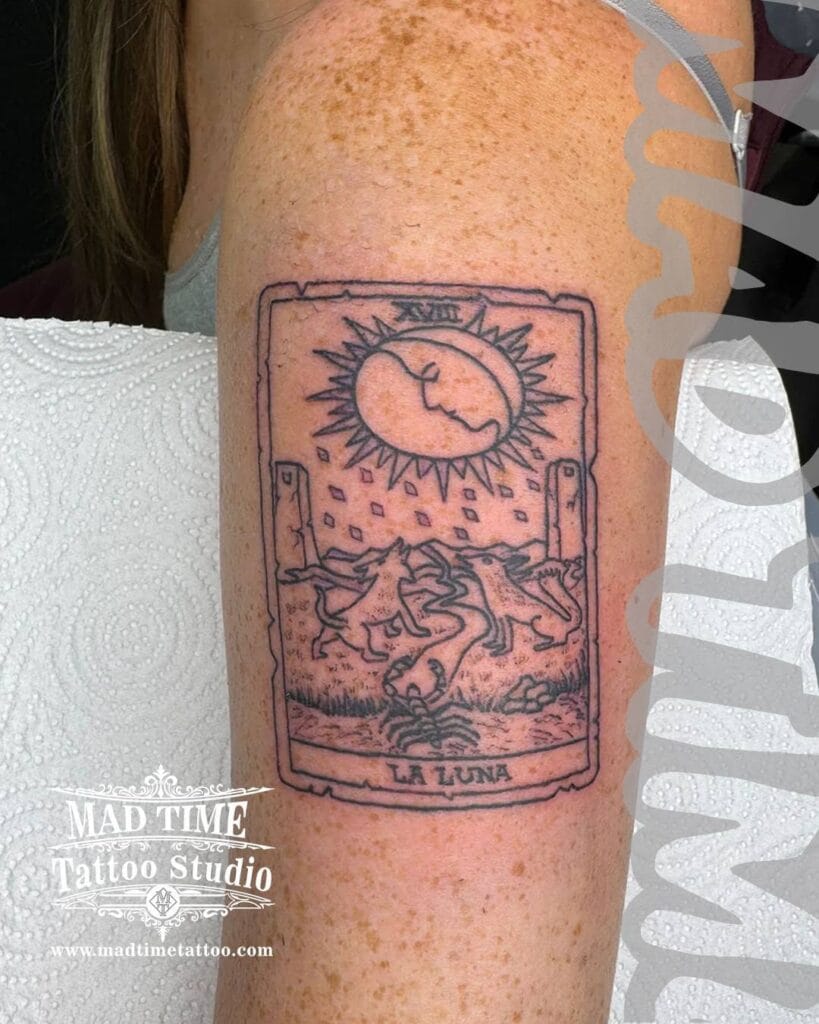 Blackwork tarot card tattoo of "La Luna" (The Moon) featuring howling wolves, a crawfish, and a crescent moon face on a woman's upper arm.