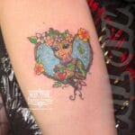 Home new dorable cartoon style tattoo of Baby Groot from Guardians of the Galaxy holding a red heart, enclosed in a floral heart frame with pink blossoms and a blue starry background, inked on an arm by Louise at Mad Time Tattoo.