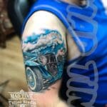 Home new Stunning color realism tattoo of a vintage blue hot rod classic car on a man's upper arm and shoulder, featuring realistic clouds, chrome reflections, and smoke effects by Mad Time Tattoo.
