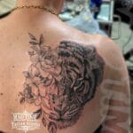 Home new Stunning black and grey realism cover-up tattoo on a woman's shoulder blade and upper back by Mad Time Tattoo, featuring a detailed tiger head with a bright blue eye hidden among blooming flowers to cover an old tribal design.