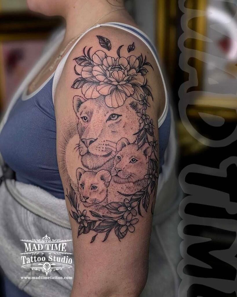 Detailed dotwork tattoo of a lioness with two small cubs surrounded by beautiful blooming flowers on a woman's upper arm and shoulder.