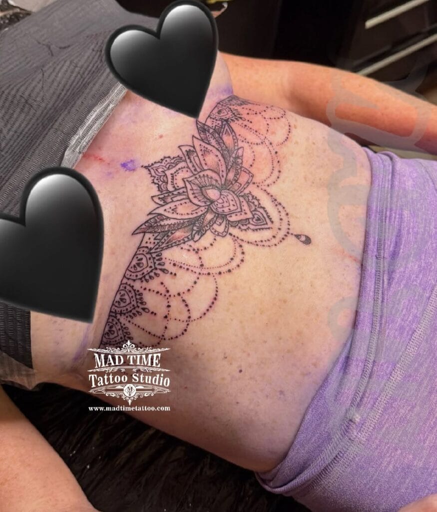Delicate fine line and dotwork underbust tattoo on a woman featuring a central ornamental lotus flower with dangling chandelier bead strings across the sternum and ribs.