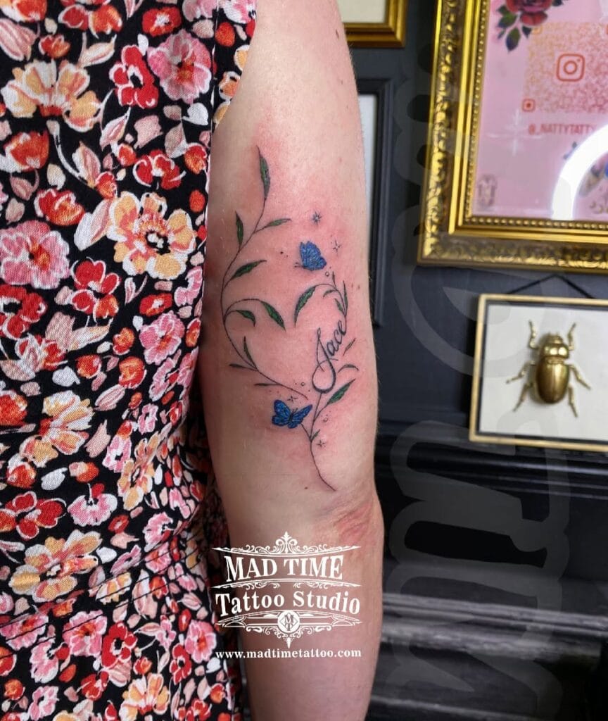 Soft color fine line floral tattoo on an arm with blue, purple, and green watercolor accents.