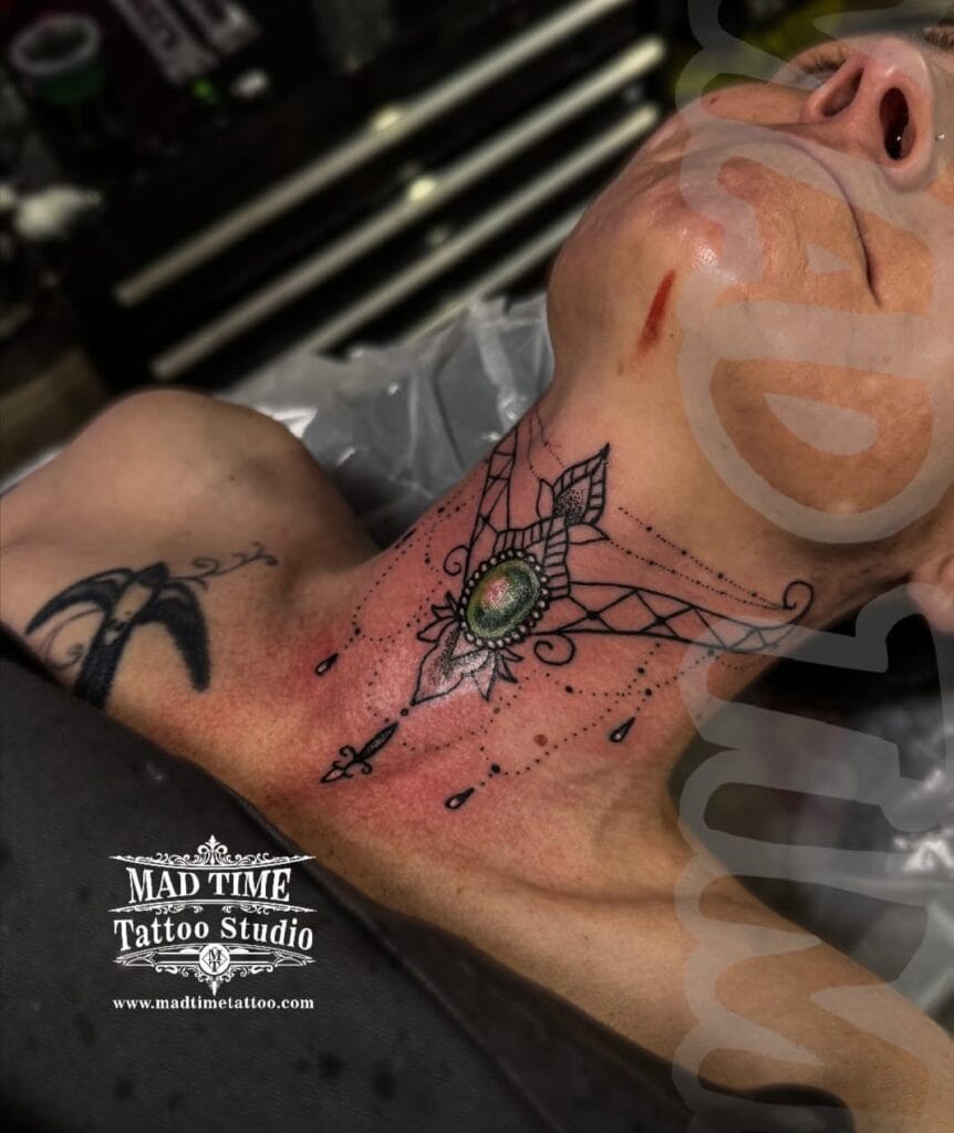 Elegant fine line ornamental neck tattoo on a woman featuring a central green gem mandala and dangling chandelier lace details on the throat.