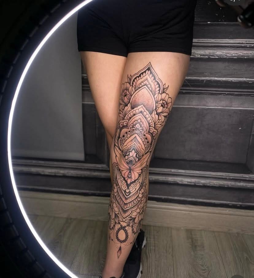 Massive black and grey ornamental tattoo covering a woman's entire front leg from thigh to ankle, featuring layered geometric mandalas, shaded flowers, and a small Taurus zodiac symbol.