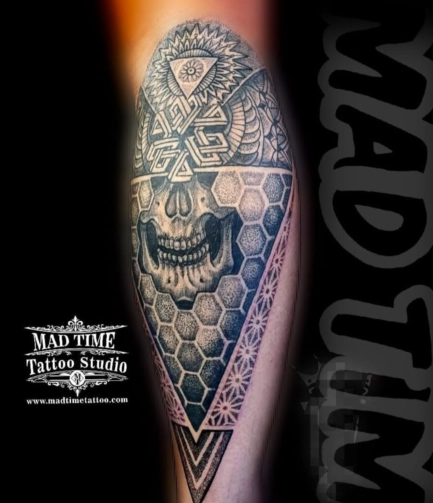 Black and grey geometric tattoo featuring a detailed human skull integrated with an All-Seeing Eye triangle, impossible geometric shapes, and a honeycomb hexagon pattern on an arm.