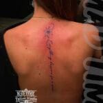 Home new Delicate fine line script tattoo on a woman's spine reading "Everything happens for a reason" flowing vertically from a minimalist lotus flower on the upper back by Mad Time Tattoo.