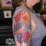 Home new Stunning watercolour style upper arm sleeve tattoo by Natalie at Mad Time Tattoo, featuring a retro vintage microphone wrapped in a "Never Can Tell" banner script, surrounded by a realistic sunflower, red roses, and colourful blue and purple watercolour paint splatters with music notes.