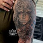 Home new Powerful black and grey realism tattoo of a Viking Valkyrie shield-maiden with piercing red eyes and an ornate Norse helmet, blended with a roaring Fenrir wolf and a Kolovrat sunwheel shield on a man's upper arm by Mad Time Tattoo.