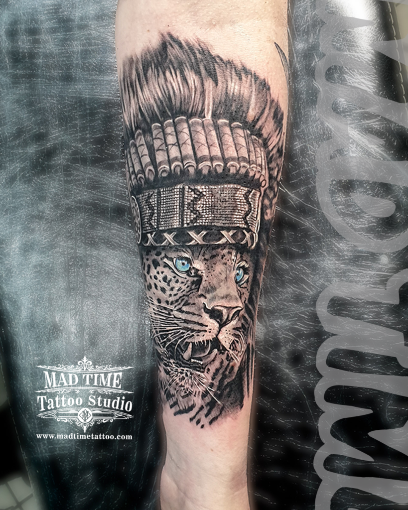 Detailed black and grey realistic jaguar tattoo wearing an intricate Indian warrior headdress with bright blue eyes by Henry at our studio
