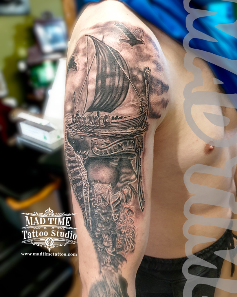 Black and grey realism shoulder tattoo of a Viking drakkar longship sailing in stormy waters above an engraved Norse warrior helmet by Henry at our studio
