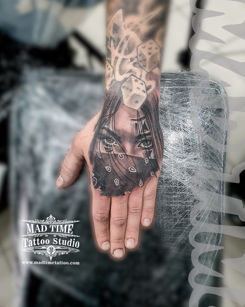 Chicano style realism hand tattoo showcasing a girl's eyes behind a paisley bandana paired with flying lucky dice on the wrist by Henry at our studio