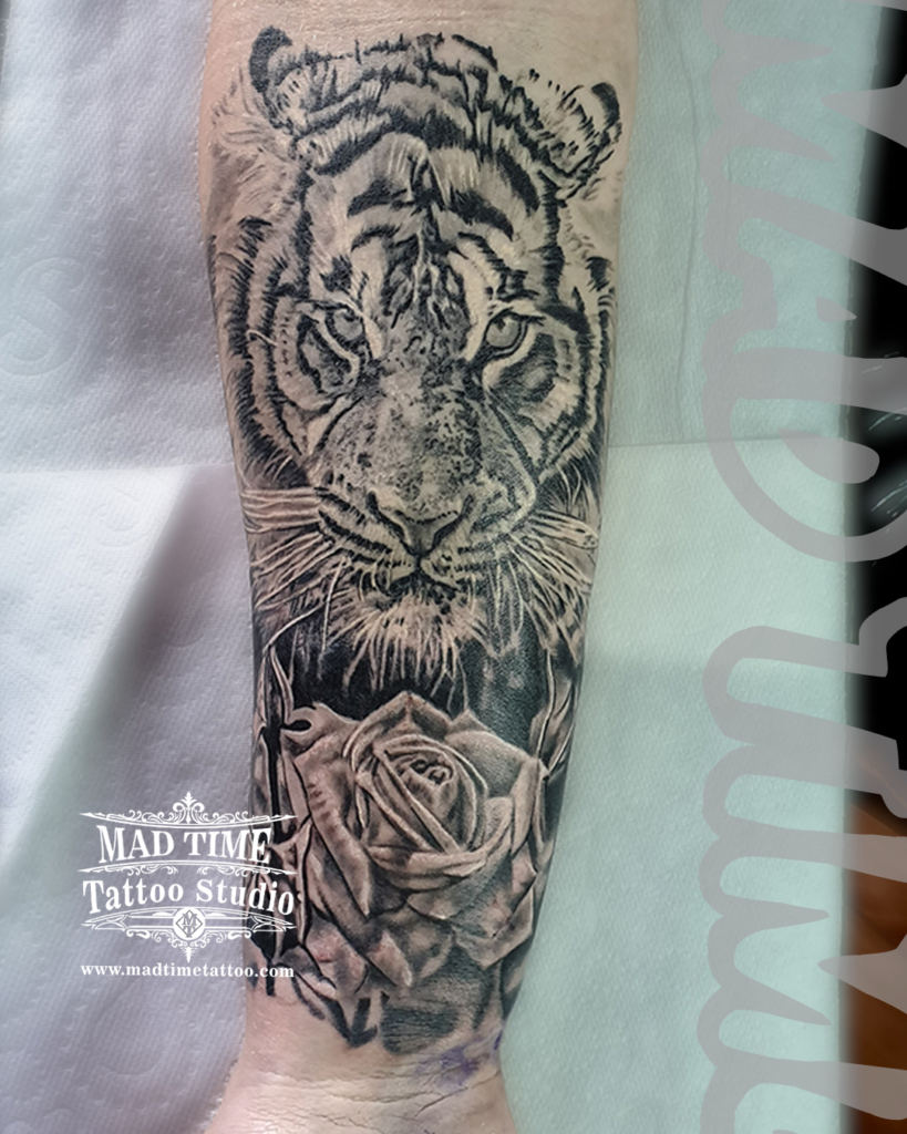 High contrast black and grey realism tattoo of an intense Bengal tiger face paired with a soft petaled rose on the wrist by Henry at our studio