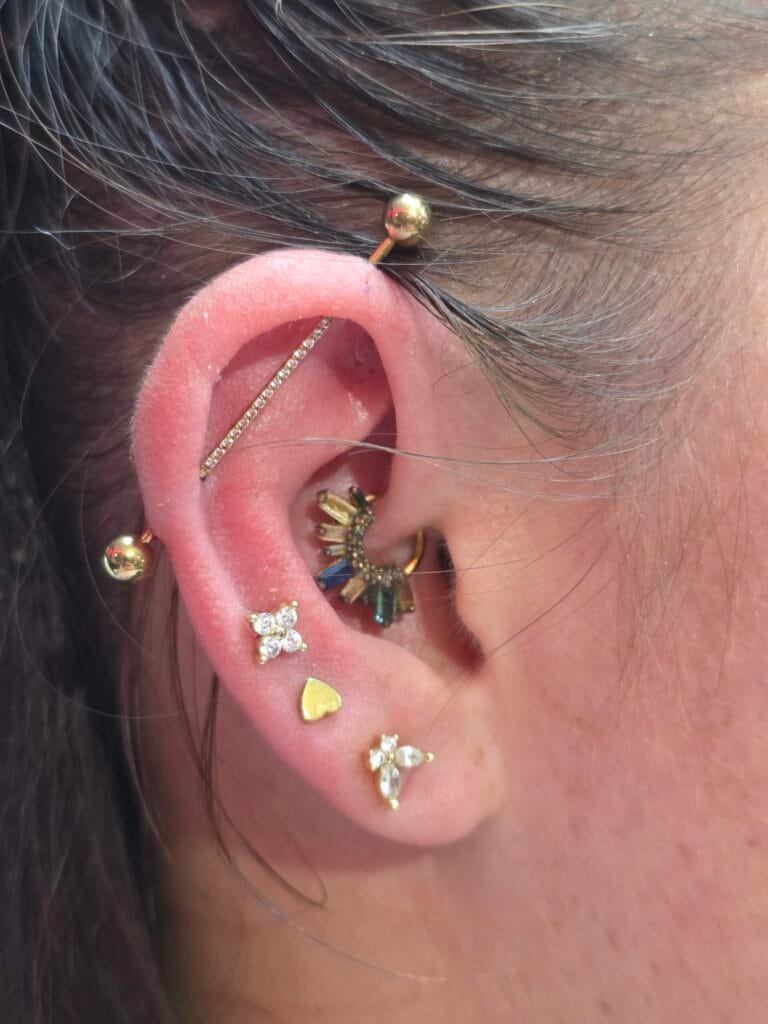 Gold industrial piercing barbell adorned with tiny pave crystals paired with a daith hoop and triple lobe earrings in a curated ear