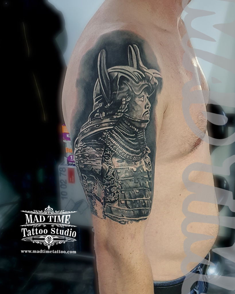 Black and grey realism shoulder tattoo of a Japanese samurai warrior in full ornate traditional armor and helmet by Henry at our studio