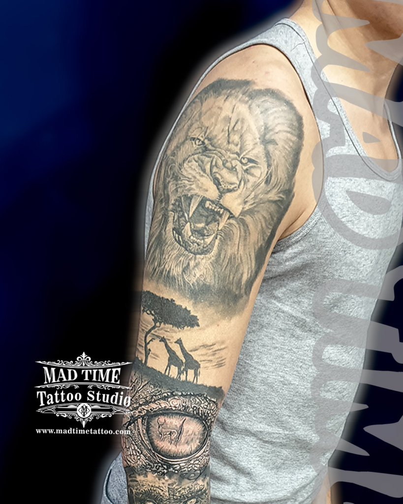 Complete black and grey realism arm sleeve tattoo featuring a roaring lion giraffes at a savanna tree and a realistic animal eye by resident artist Henry at Mad Time Tattoo Studio