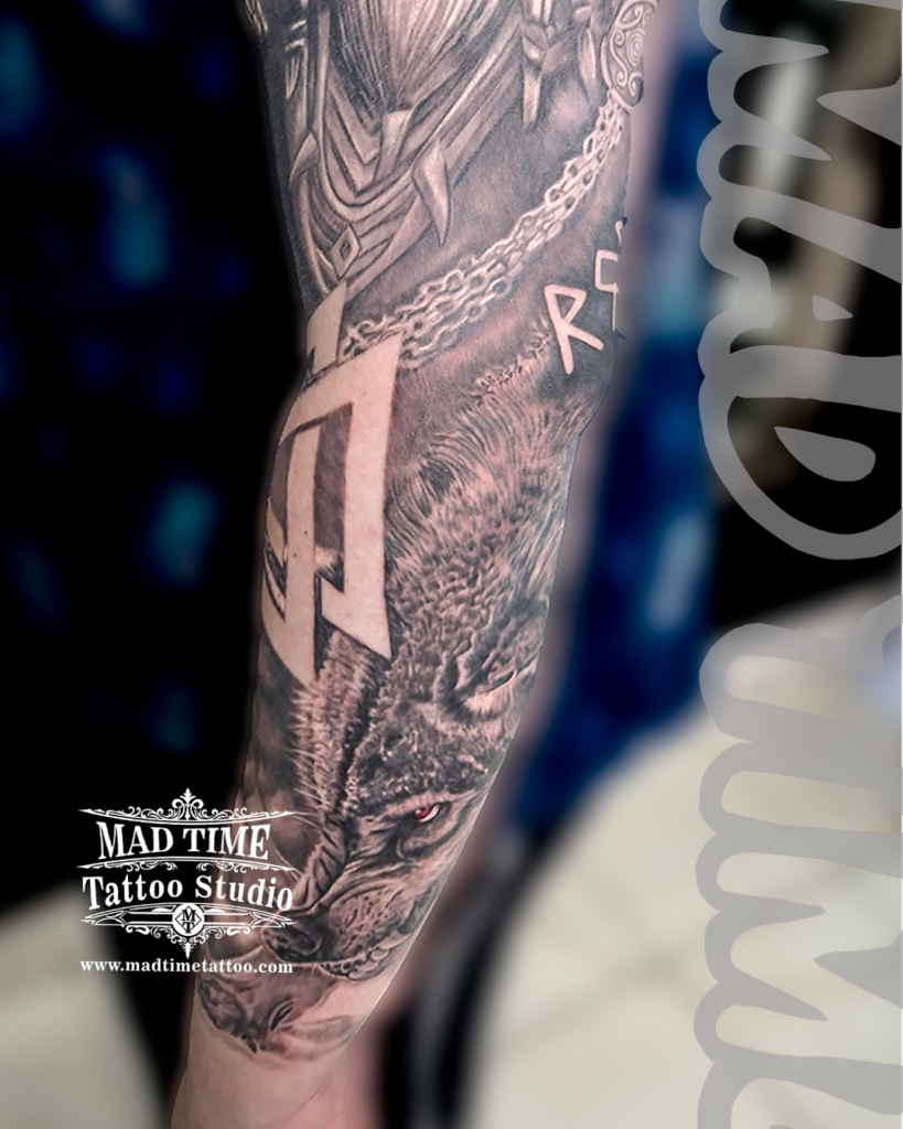 Black and grey realism forearm tattoo combining engraved Norse Viking armor scales runic letters and a wild wolf portrait with a custom red eye accent by Henry at Mad Time Tattoo Studio