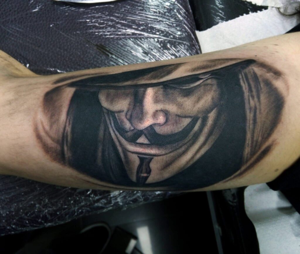 Realistic black and grey Anonymous hacker mask tattoo with smooth shading and deep black background by guest artist Pablo from Spain at our studio