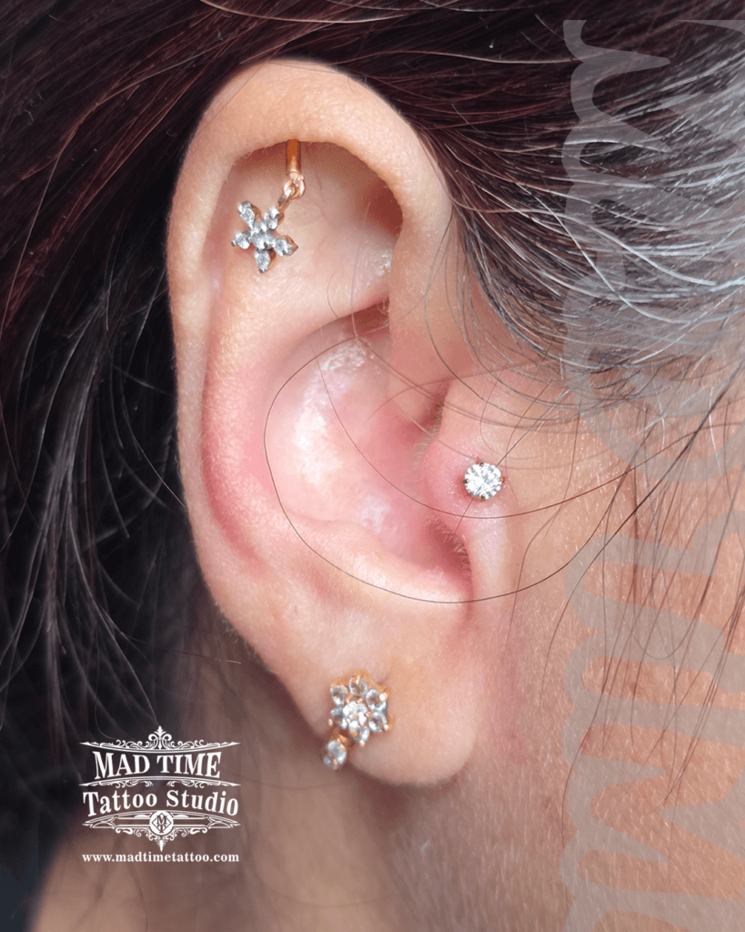 Gold helix piercing with a dangling floral charm set with crystals paired with a gold tragus stud at Mad Time Tattoo Studio
