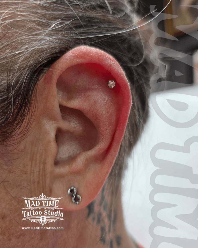 Minimalist single helix piercing with a tiny clear crystal stud and an infinity sign earring in the lobe at Mad Time Tattoo Studio
