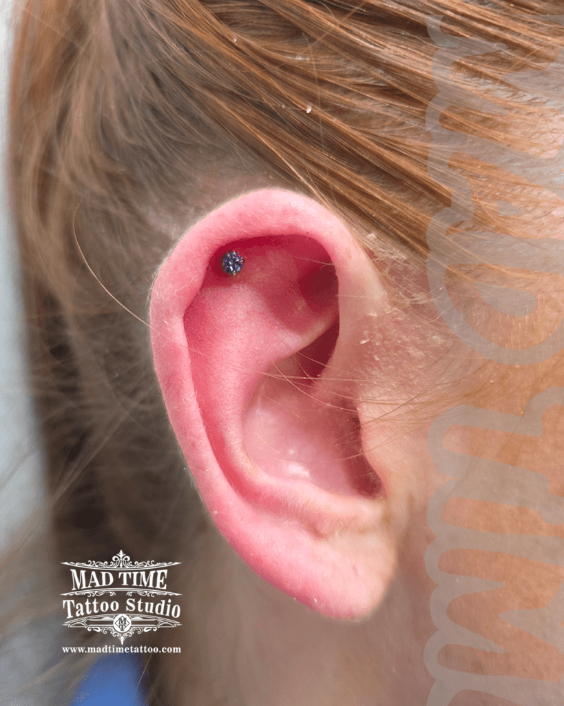 Flat helix piercing with a prong-set dark violet crystal stud earring in the upper ear cartilage at Mad Time Tattoo Studio