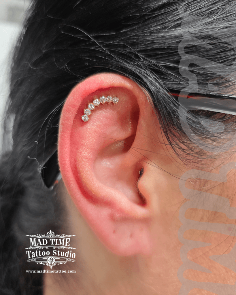 Elegant gold helix piercing with a seven-stone crystal cluster attachment contouring the ear rim at Mad Time Tattoo Studio
