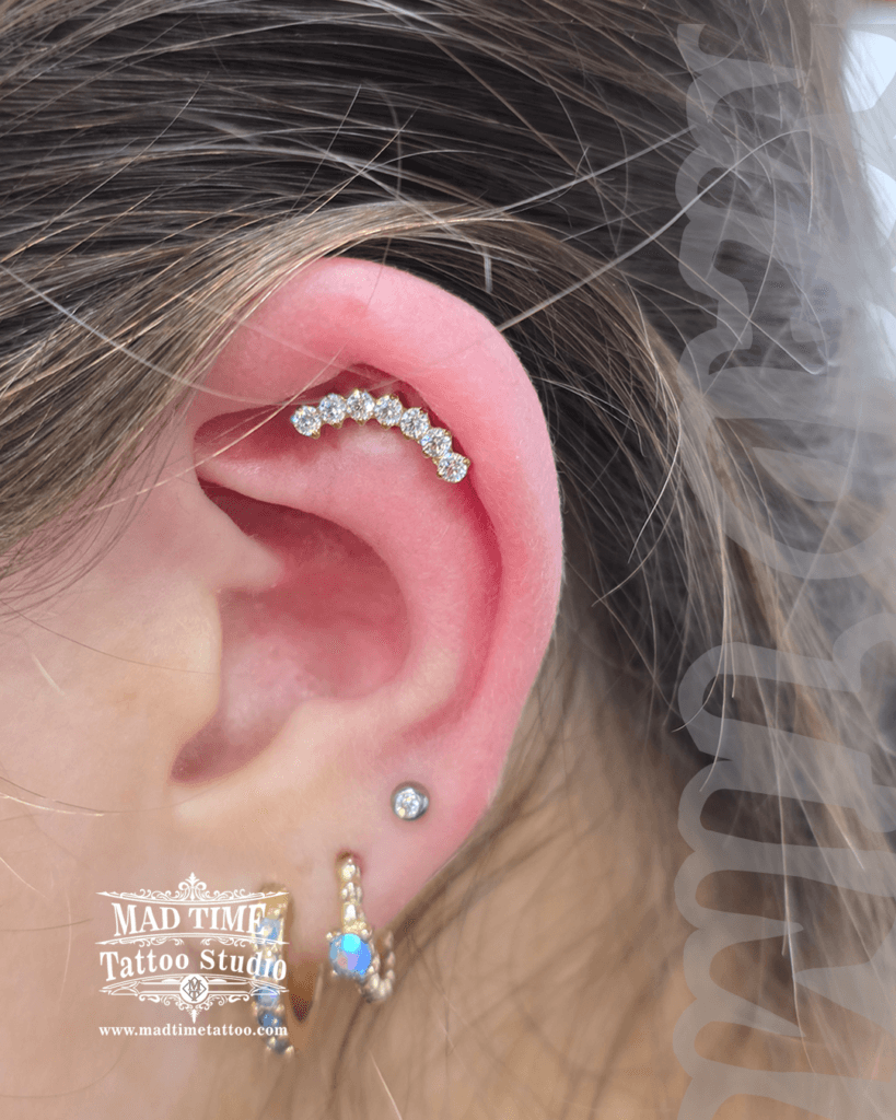 Curved gold helix piercing barbell featuring a row of seven pave crystals and textured gold hoop earrings with an opal stone at Mad Time Tattoo Studio