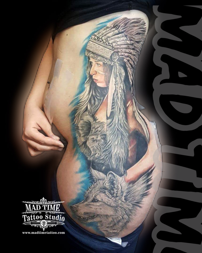 Large scale side ribs and torso black and grey realism tattoo of a Native American woman in a full feathered headdress and a wild wolf by our studio